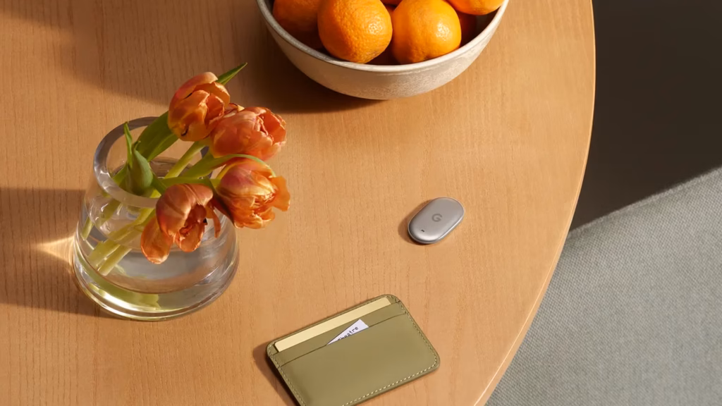 Google lança pixel watch 5 e pixel tag no made by google 2026 Pixel tag prateado ao lado de uma carteira verde sobre mesa de madeira, com vaso de tulipas e tigela de laranjas ao fundo.